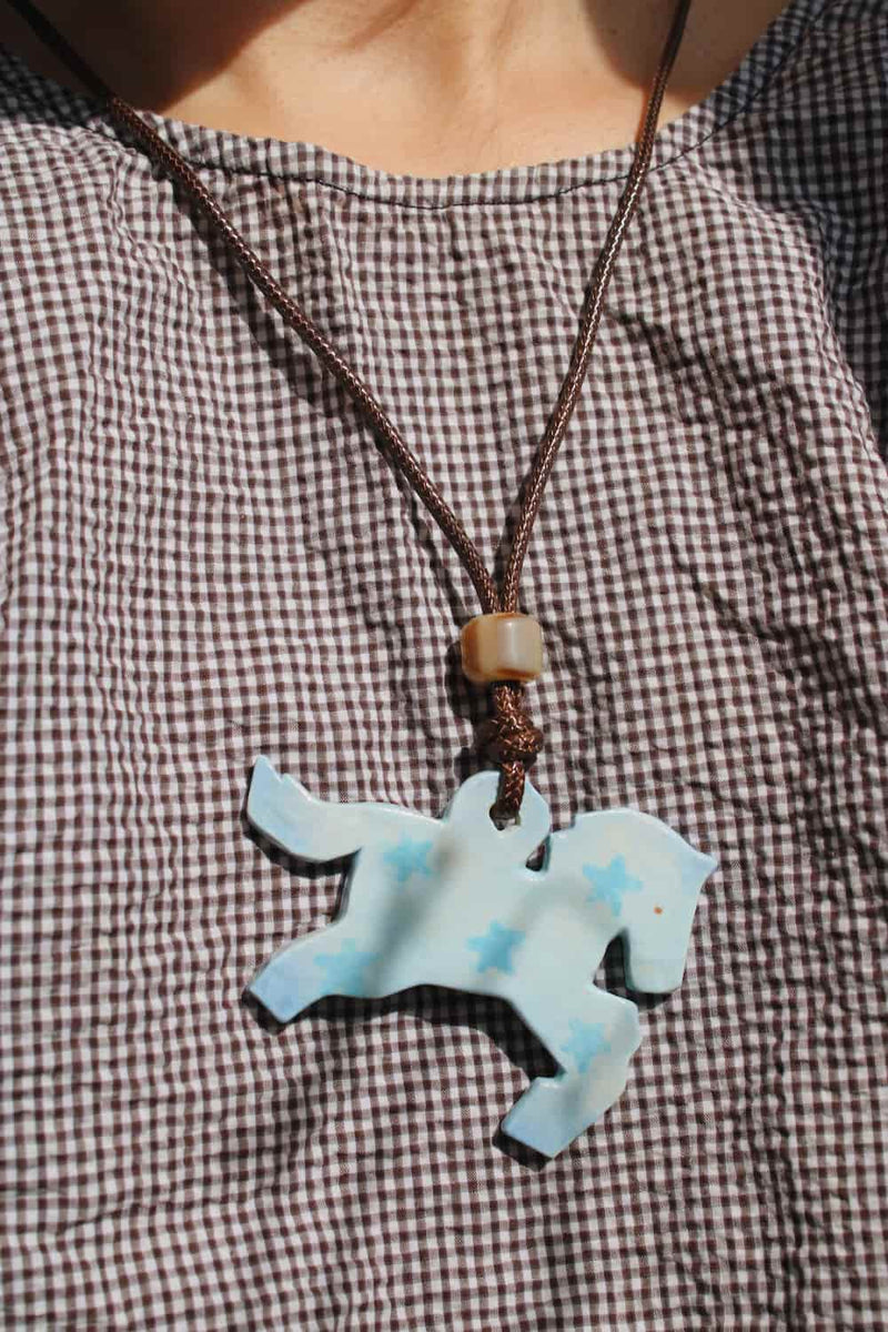 Stardust Pony Charm Necklace in pale blue with star detail worn on a brown gingham top in natural sunlight.