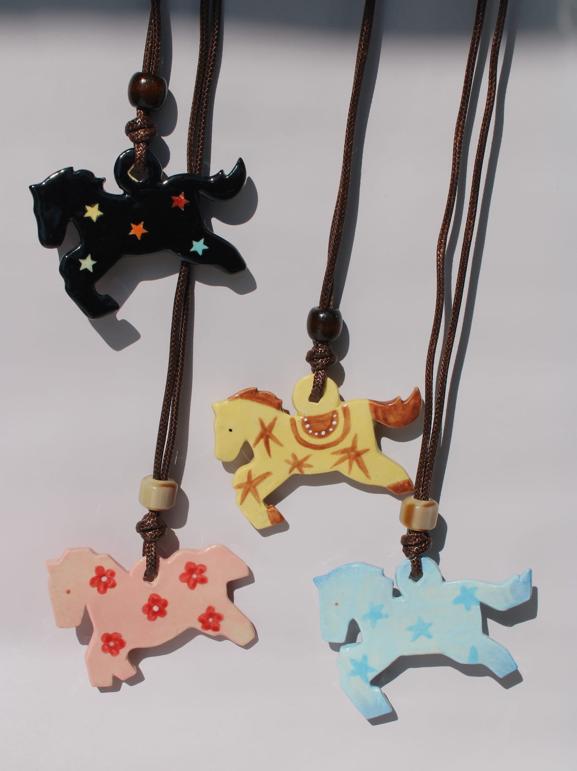 Pony Charm Necklace collection with four ceramic horse pendant designs — black starry night, yellow saddle pattern, pink floral Petal Pony, and blue starry Stardust Pony — on brown cords