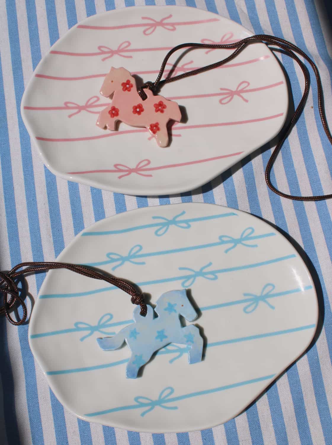Petal Pony and Stardust Pony ceramic charm necklaces displayed together on a blue striped surface, featuring pink floral and pale blue bow motifs on leather cords
