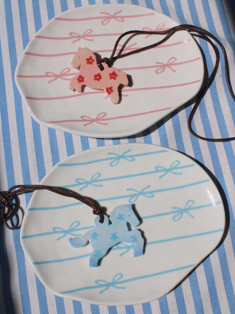 Petal Pony and Stardust Pony ceramic charm necklaces displayed together on a blue striped surface, featuring pink floral and pale blue bow motifs on leather cords