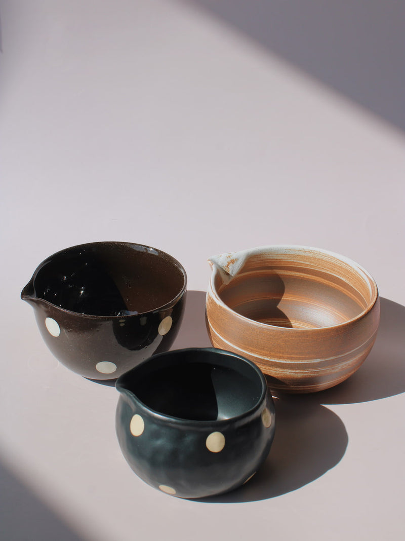 Kanso Matcha Bowl, Mochi Ceramic Matcha Bowl, and Amber Matcha Bowl displayed together — featuring a terracotta swirl glaze, a glossy black with white dot pattern, and a matte teal with white dot pattern — each with a pouring spout, on a soft pink background.