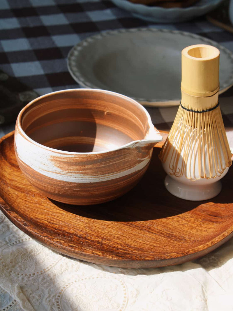 Kanso Matcha Bowl in warm terracotta with white swirl glaze, filled with a matcha latte with milk swirl, styled on a wooden tray with a bamboo chasen whisk and chashaku scoop, on a lace cloth with a dark gingham surface in the background.