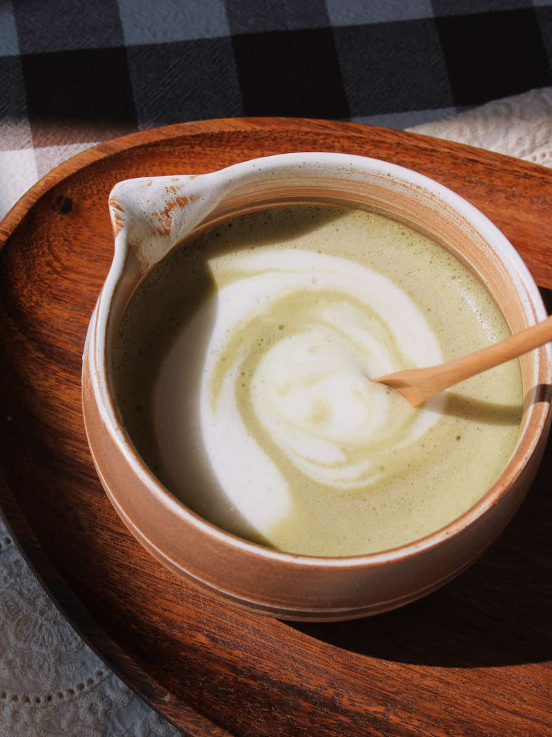 Kanso Matcha Bowl in warm terracotta with white swirl glaze, filled with a matcha latte with milk swirl, styled on a wooden tray with a bamboo chasen whisk and chashaku scoop, on a lace cloth with a dark gingham surface in the background.