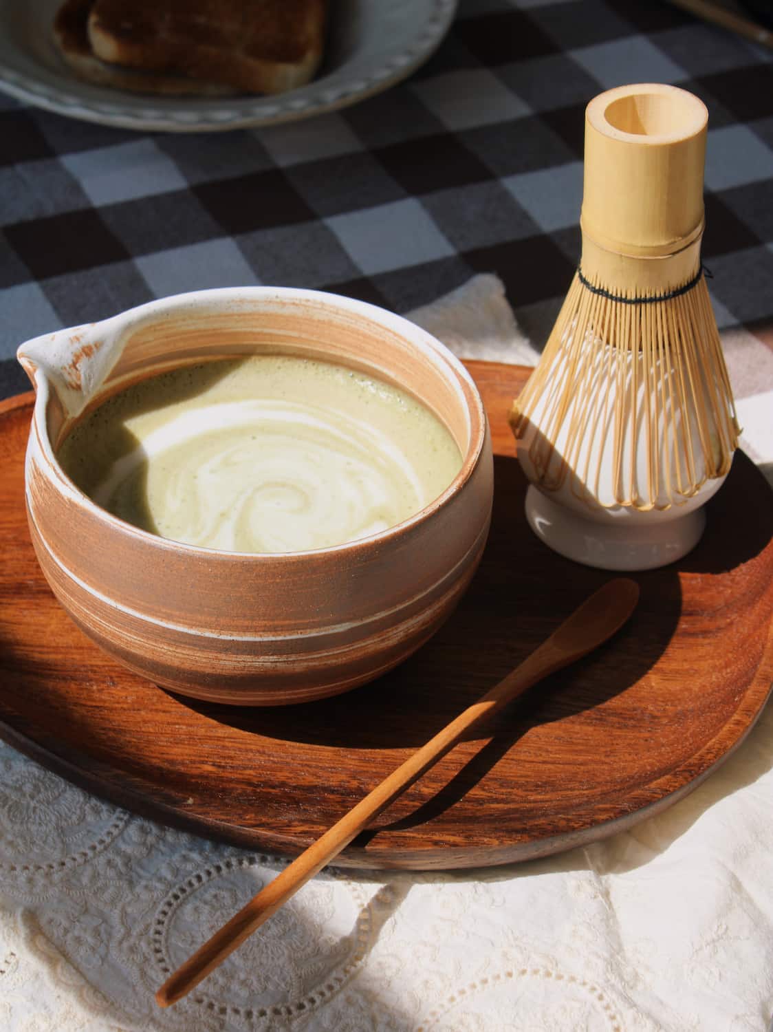 Kanso Matcha Bowl in warm terracotta with white swirl glaze, filled with a matcha latte with milk swirl, styled on a wooden tray with a bamboo chasen whisk and chashaku scoop, on a lace cloth with a dark gingham surface in the background.