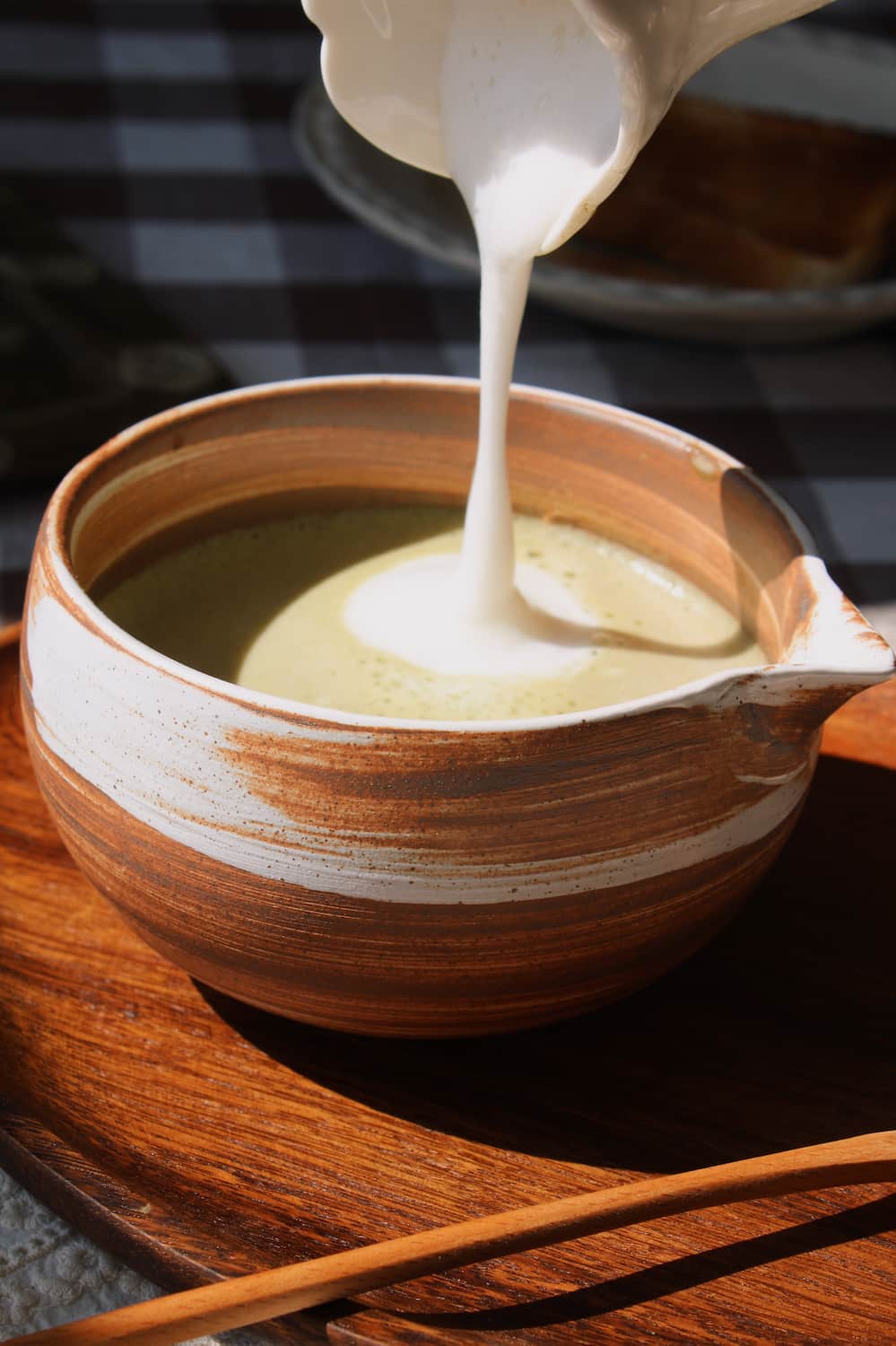Kanso Matcha Bowl in warm terracotta with white swirl glaze, filled with a matcha latte with milk swirl, styled on a wooden tray with a bamboo chasen whisk and chashaku scoop, on a lace cloth with a dark gingham surface in the background.