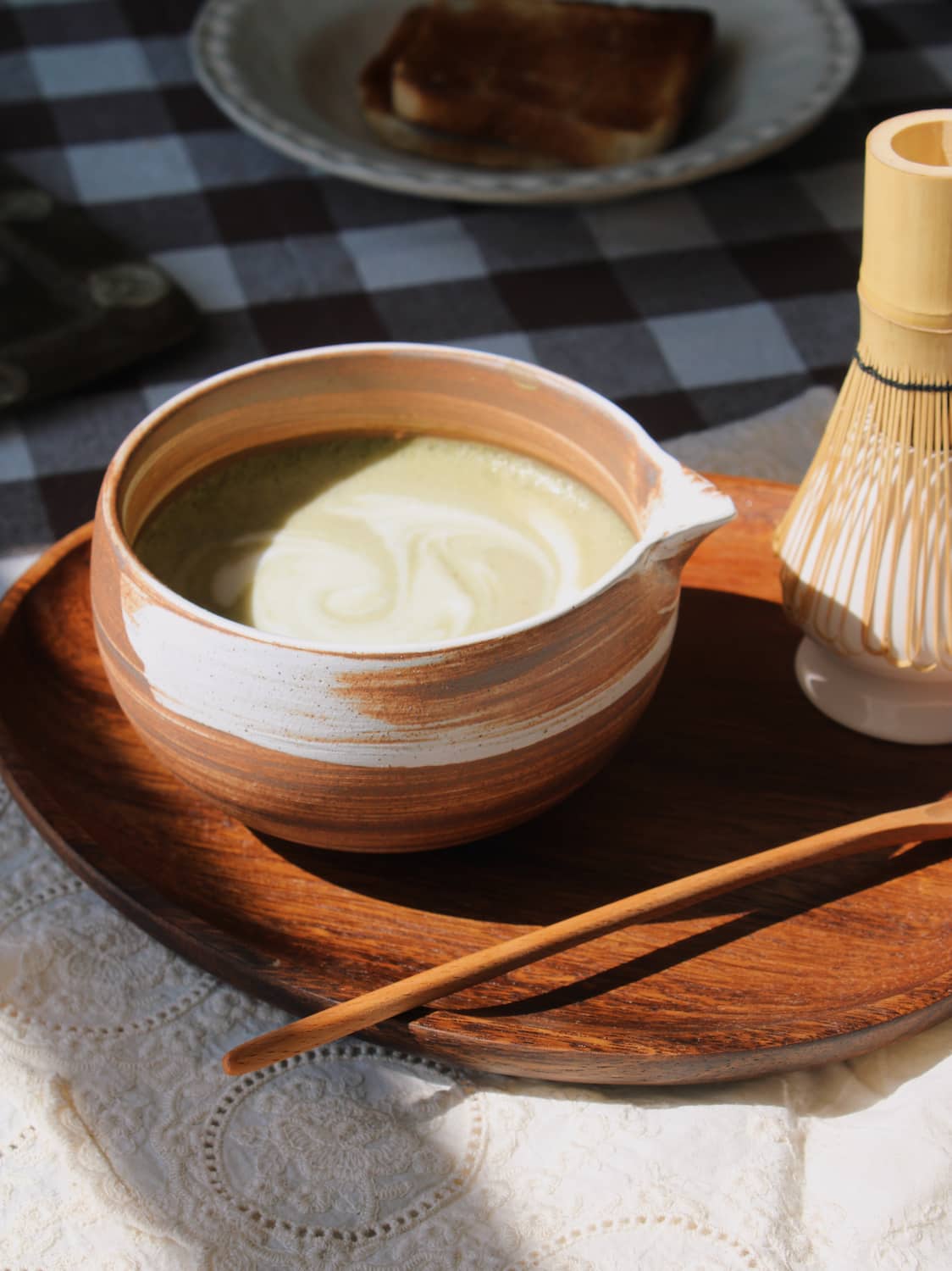 Kanso Matcha Bowl in warm terracotta with white swirl glaze, filled with a matcha latte with milk swirl, styled on a wooden tray with a bamboo chasen whisk and chashaku scoop, on a lace cloth with a dark gingham surface in the background.