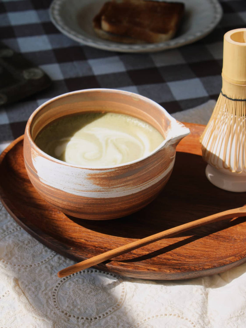 Kanso Matcha Bowl in warm terracotta with white swirl glaze, filled with a matcha latte with milk swirl, styled on a wooden tray with a bamboo chasen whisk and chashaku scoop, on a lace cloth with a dark gingham surface in the background.