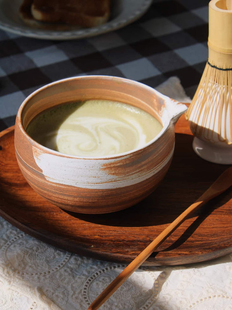 Kanso Matcha Bowl in warm terracotta with white swirl glaze, filled with a matcha latte with milk swirl, styled on a wooden tray with a bamboo chasen whisk and chashaku scoop, on a lace cloth with a dark gingham surface in the background.