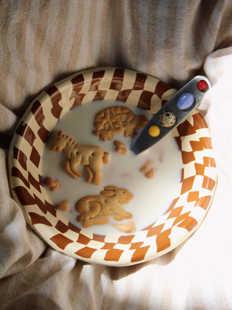 Nova Ceramic Bowl in cream with brown checkerboard pattern, filled with milk and animal crackers alongside the Constella Spoon with colourful gemstone handle, on a linen surface in natural sunlight.