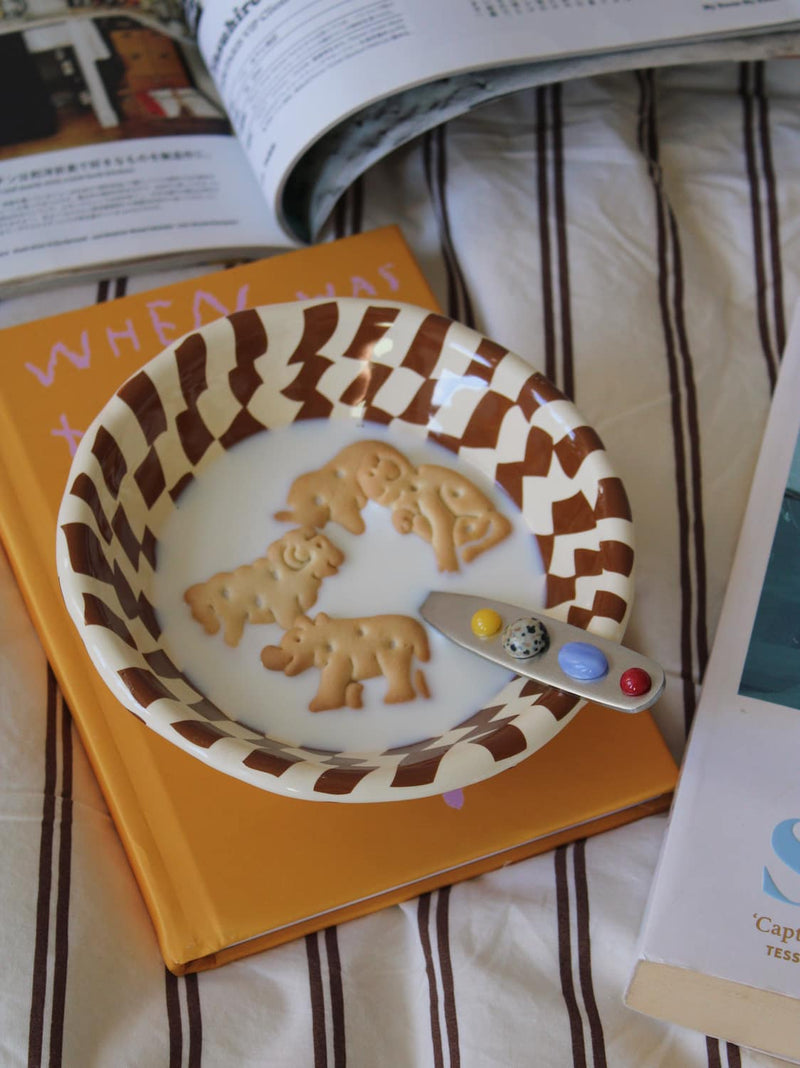 Nova Ceramic Bowl in cream with brown checkerboard pattern, filled with milk and animal crackers alongside the Constella Spoon with colourful gemstone handle, on a linen surface in natural sunlight.