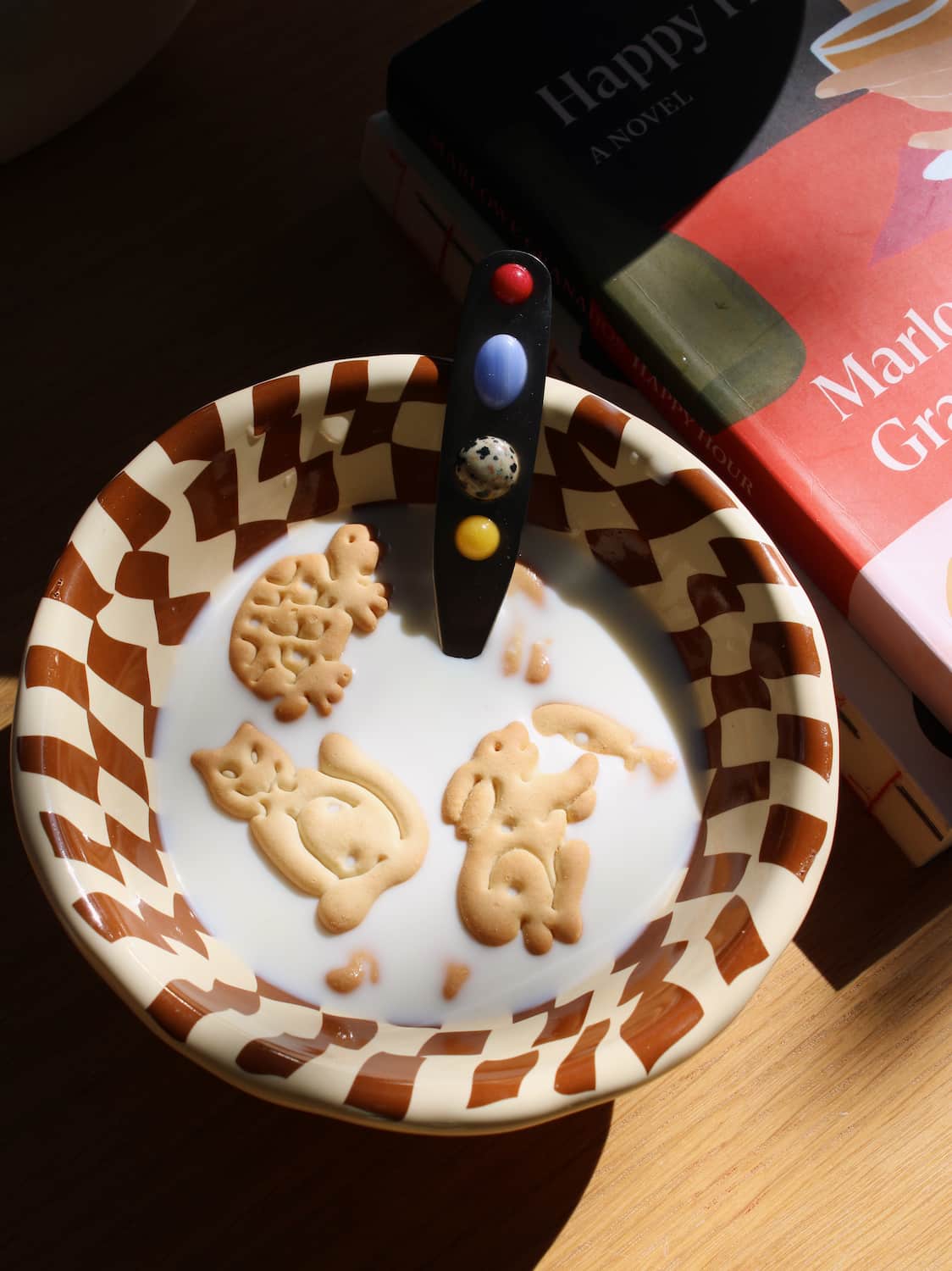 Nova Ceramic Bowl in cream with brown checkerboard pattern, filled with milk and animal crackers alongside the Constella Spoon with colourful gemstone handle, on a linen surface in natural sunlight.