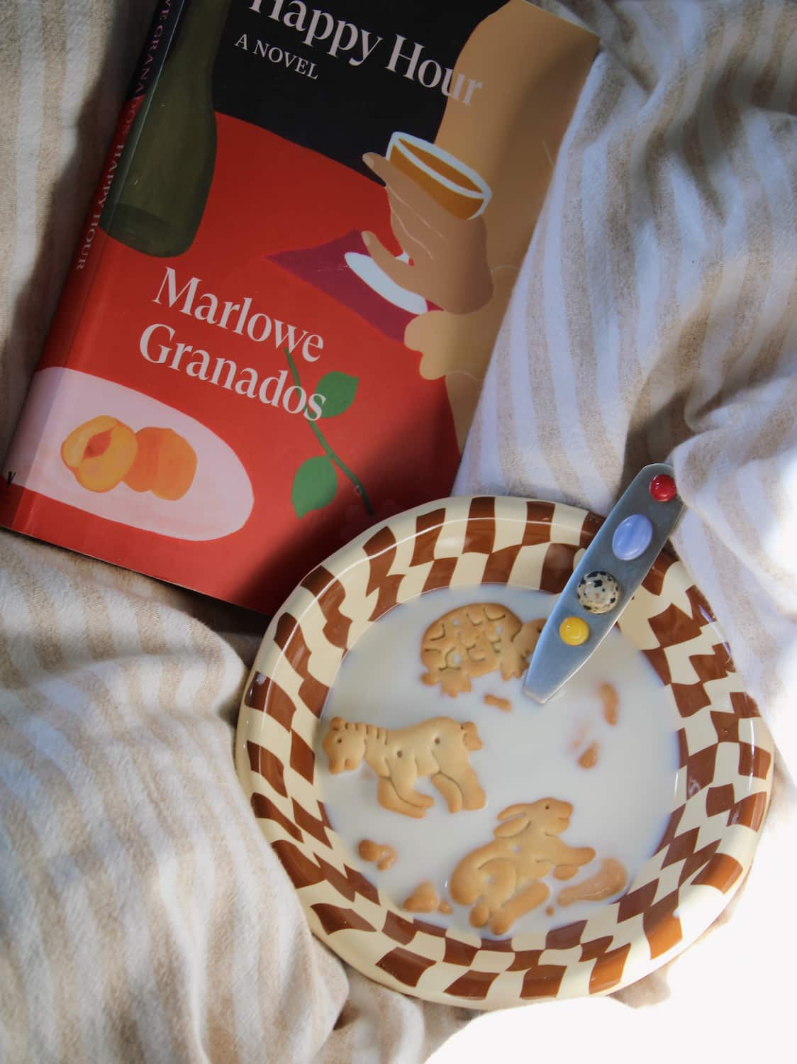 Nova Ceramic Bowl in cream with brown checkerboard pattern, filled with milk and animal crackers alongside the Constella Spoon with colourful gemstone handle, on a linen surface in natural sunlight.