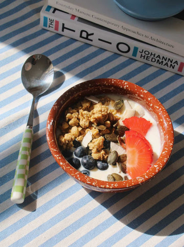 Handmade speckled ceramic bowl with yogurt, granola and fruit, styled with spoon for a relaxed breakfast table