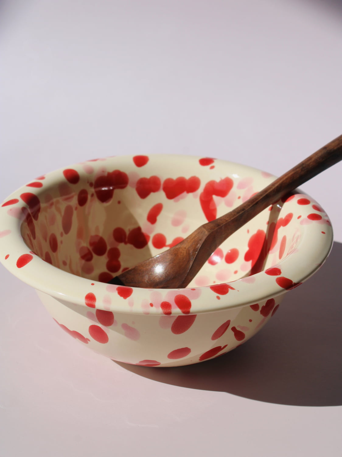 Dotty Ceramic Bowl in cream with red speckled dot pattern, paired with a wooden serving spoon, on a soft pink background.