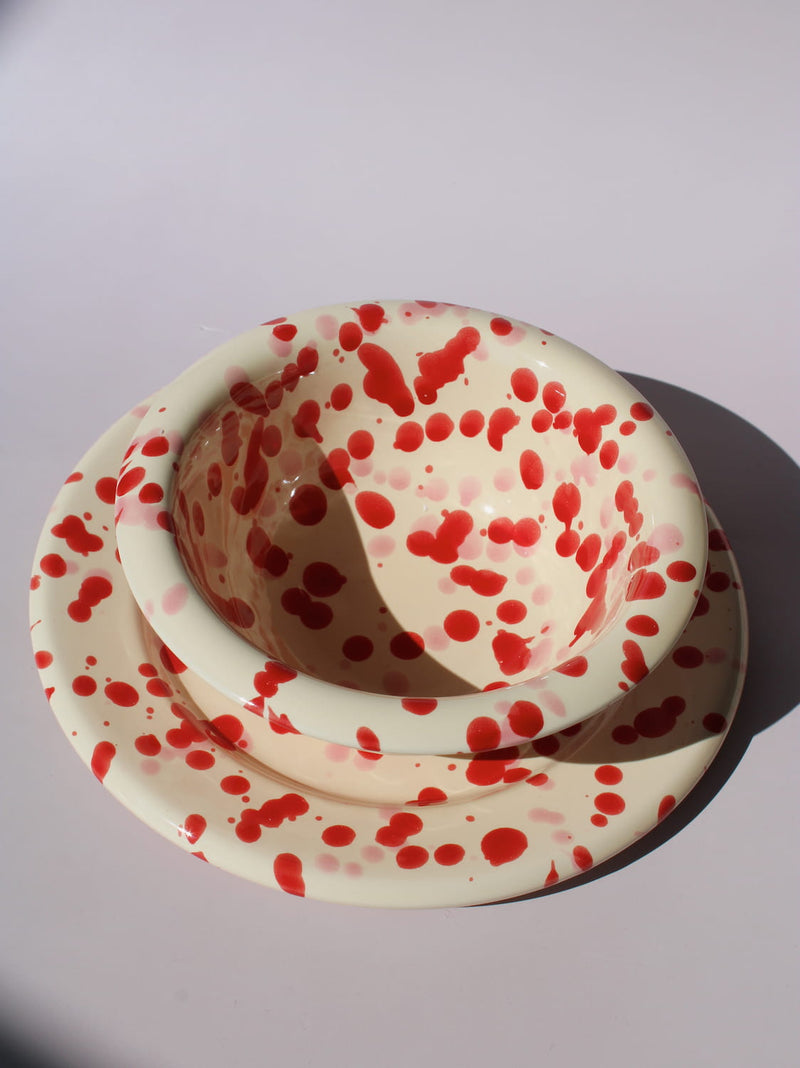 Dotty Ceramic Bowl and Dotty Ceramic Plate, a matching cream dinnerware set with red speckled dot pattern, on a soft pink background.
