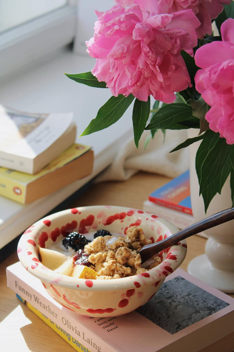 Dotty Ceramic Bowl filled with granola, blackberries and yogurt with a wooden spoon, styled on a stack of books beside pink peonies on a wooden surface.
