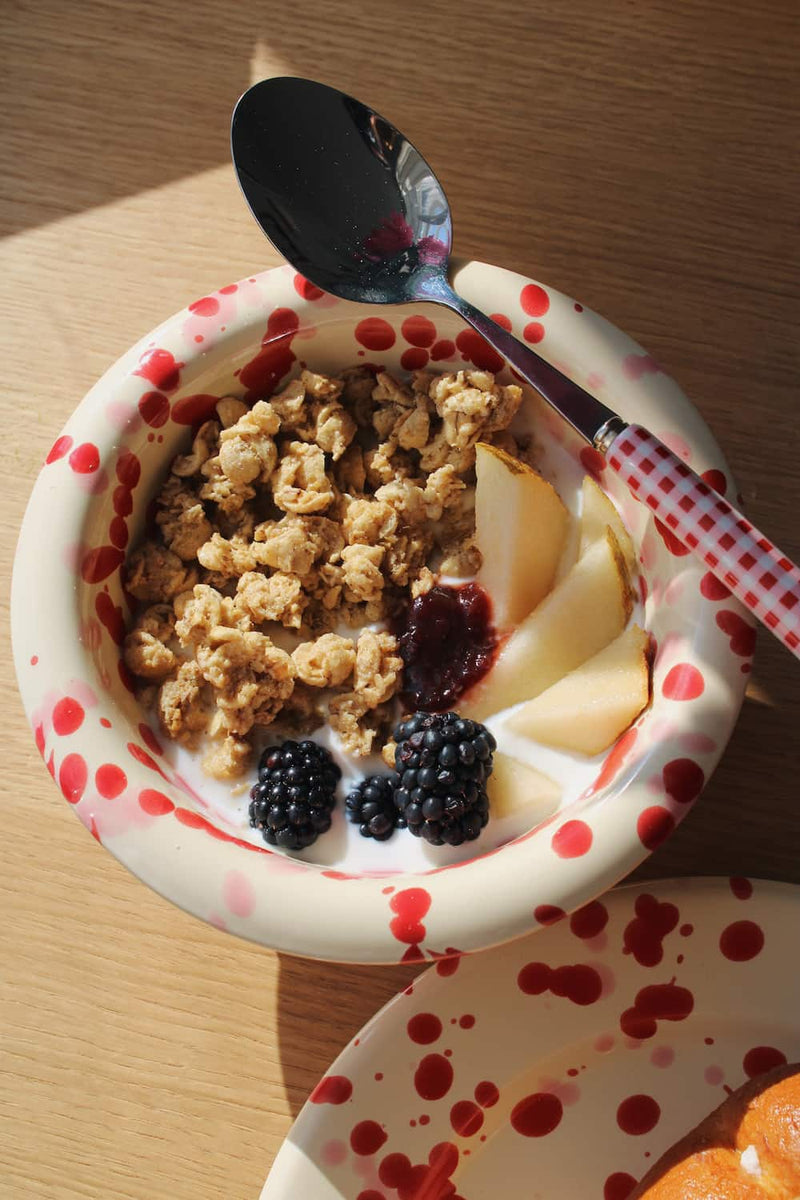 Dotty Ceramic Bowl filled with granola, blackberries and yogurt with a wooden spoon, styled on a stack of books beside pink peonies on a wooden surface.