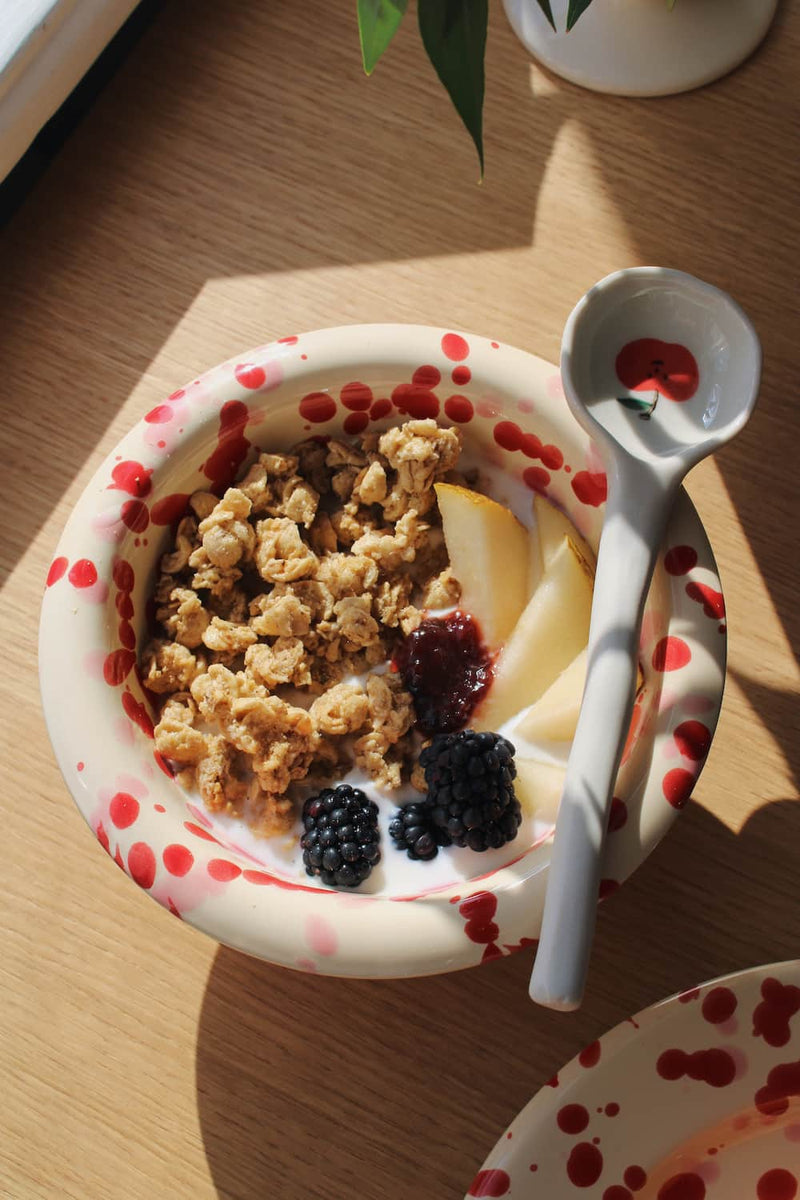 Dotty Ceramic Bowl filled with granola, blackberries and yogurt with a wooden spoon, styled on a stack of books beside pink peonies on a wooden surface.