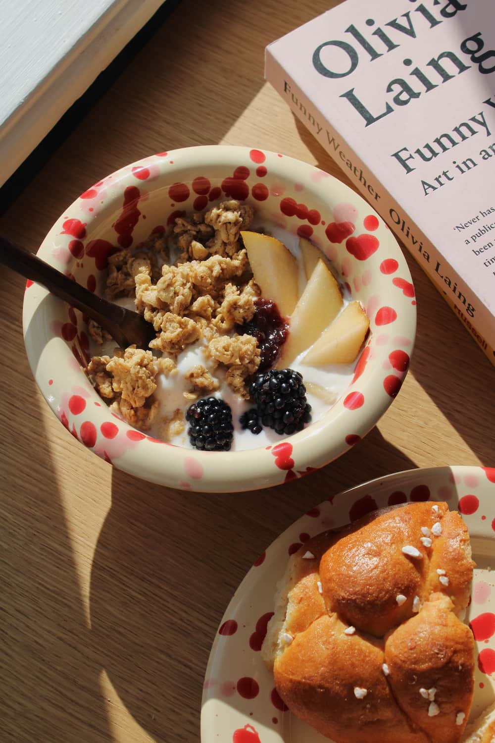 Dotty Ceramic Bowl filled with granola, blackberries and yogurt with a wooden spoon, styled on a stack of books beside pink peonies on a wooden surface.