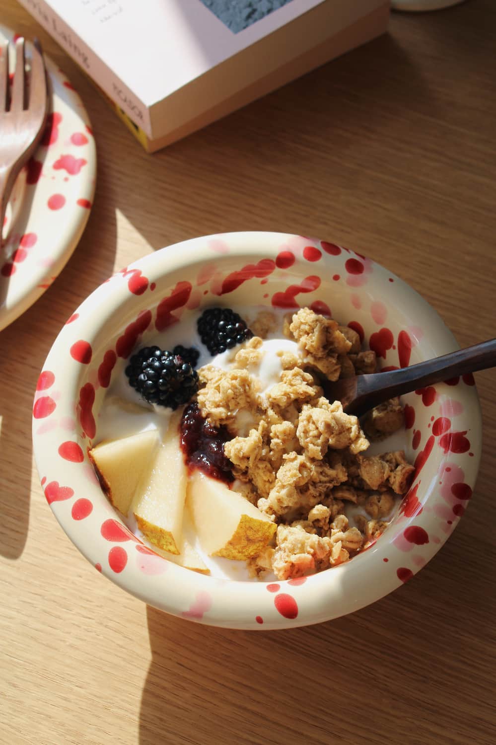 Dotty Ceramic Bowl filled with granola, blackberries and yogurt with a wooden spoon, styled on a stack of books beside pink peonies on a wooden surface.