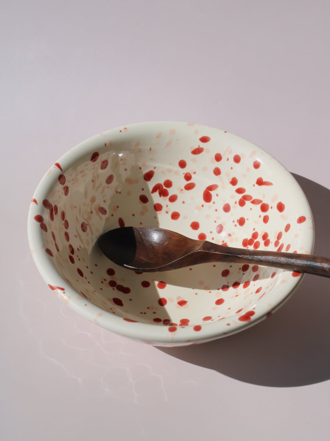 Bera Ceramic Bowl in cream with red speckled dot pattern, paired with a wooden serving spoon, on a soft pink background.