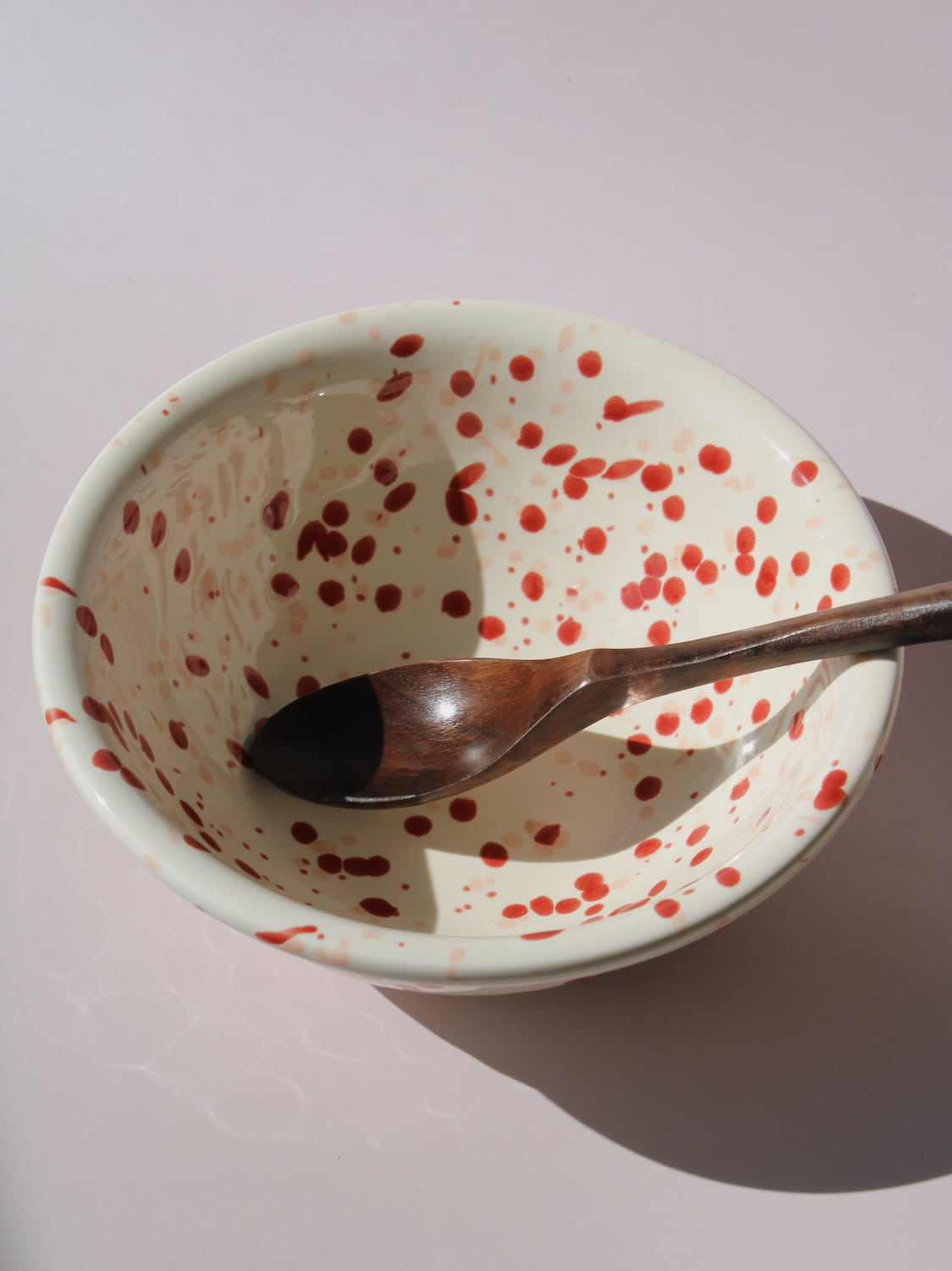 Bera Ceramic Bowl in cream with red speckled dot pattern, paired with a wooden serving spoon, on a soft pink background.
