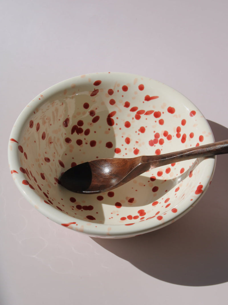 Bera Ceramic Bowl in cream with red speckled dot pattern, paired with a wooden serving spoon, on a soft pink background.