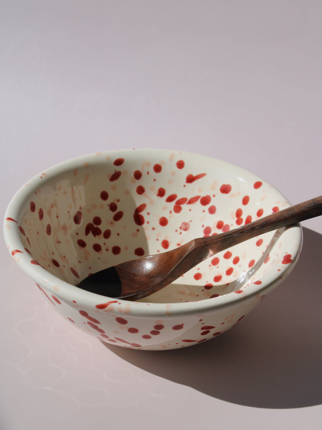 Bera Ceramic Bowl in cream with red speckled dot pattern, paired with a wooden serving spoon, on a soft pink background.
