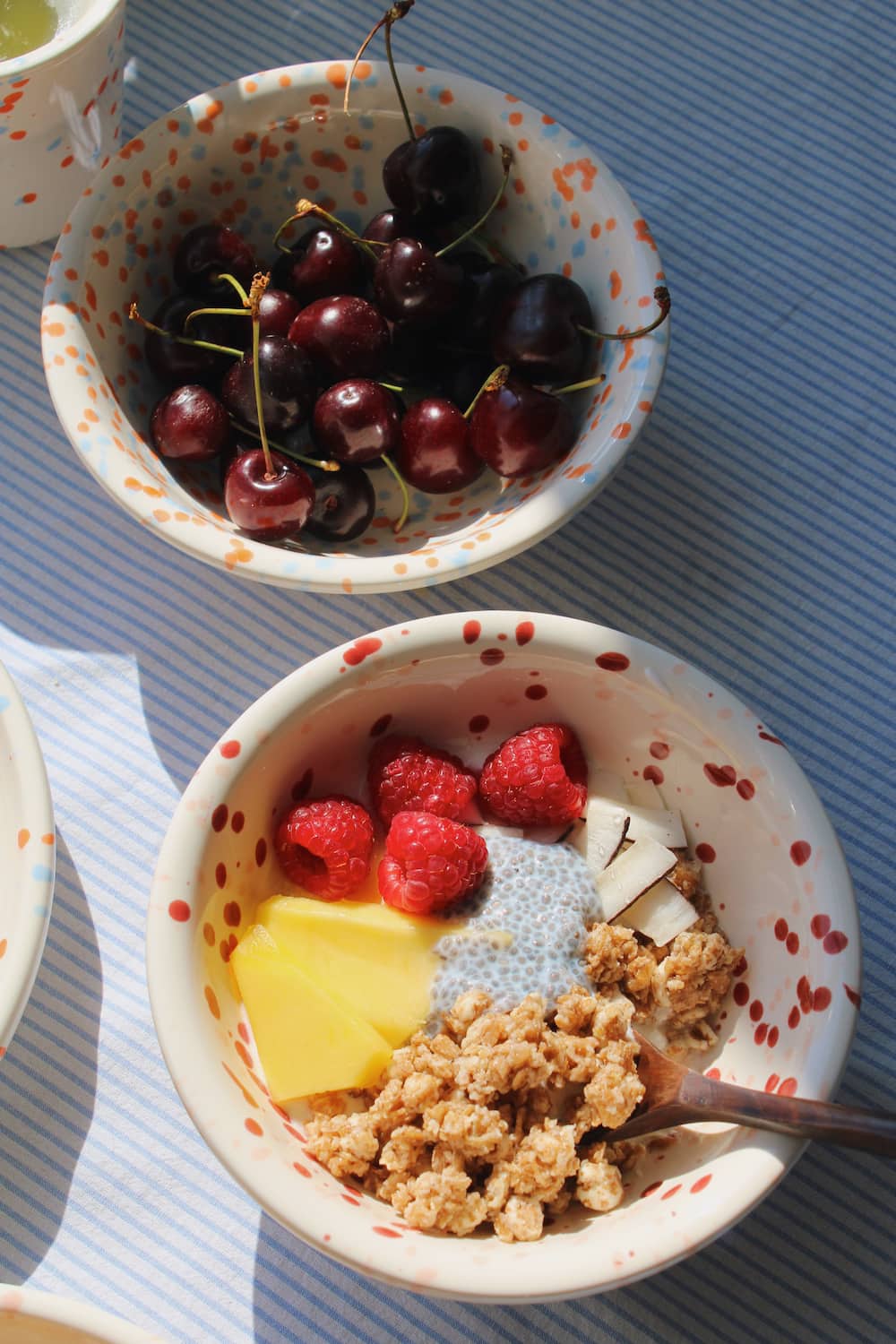 Bera Ceramic Bowl and Naro Ceramic Bowl styled together with granola, mango, raspberries and chia seeds, with the Naro Ceramic Cup visible in the background on a blue striped surface.