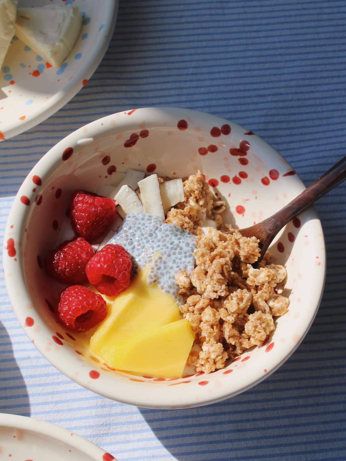 Bera Ceramic Bowl in cream with red speckled dot pattern, filled with granola, mango, raspberries and chia seeds with a wooden spoon, on a blue striped surface in natural sunlight.