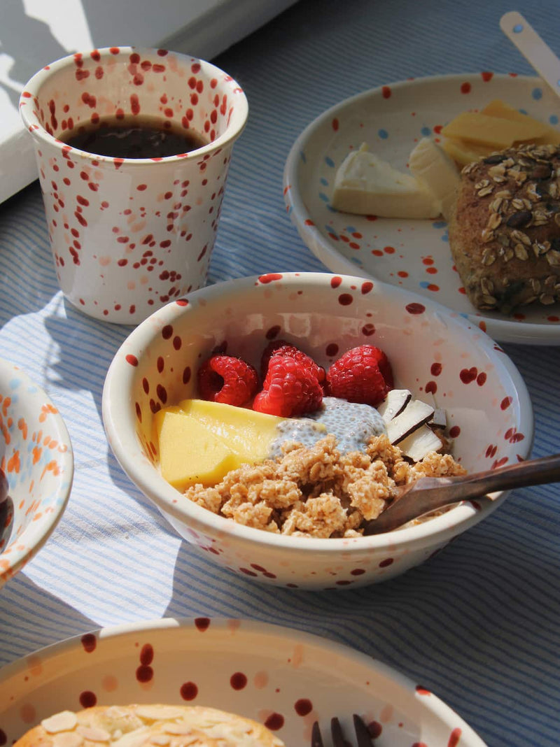 Bera Ceramic Bowl filled with granola, mango and raspberries alongside the Bera Ceramic Cup on a blue striped surface in natural sunlight.