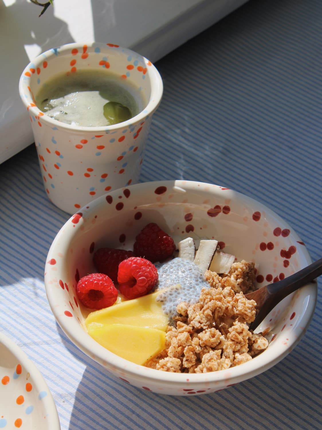 Bera Ceramic Bowl filled with granola, mango and raspberries alongside the Naro Ceramic Cup on a blue striped surface in natural sunlight.