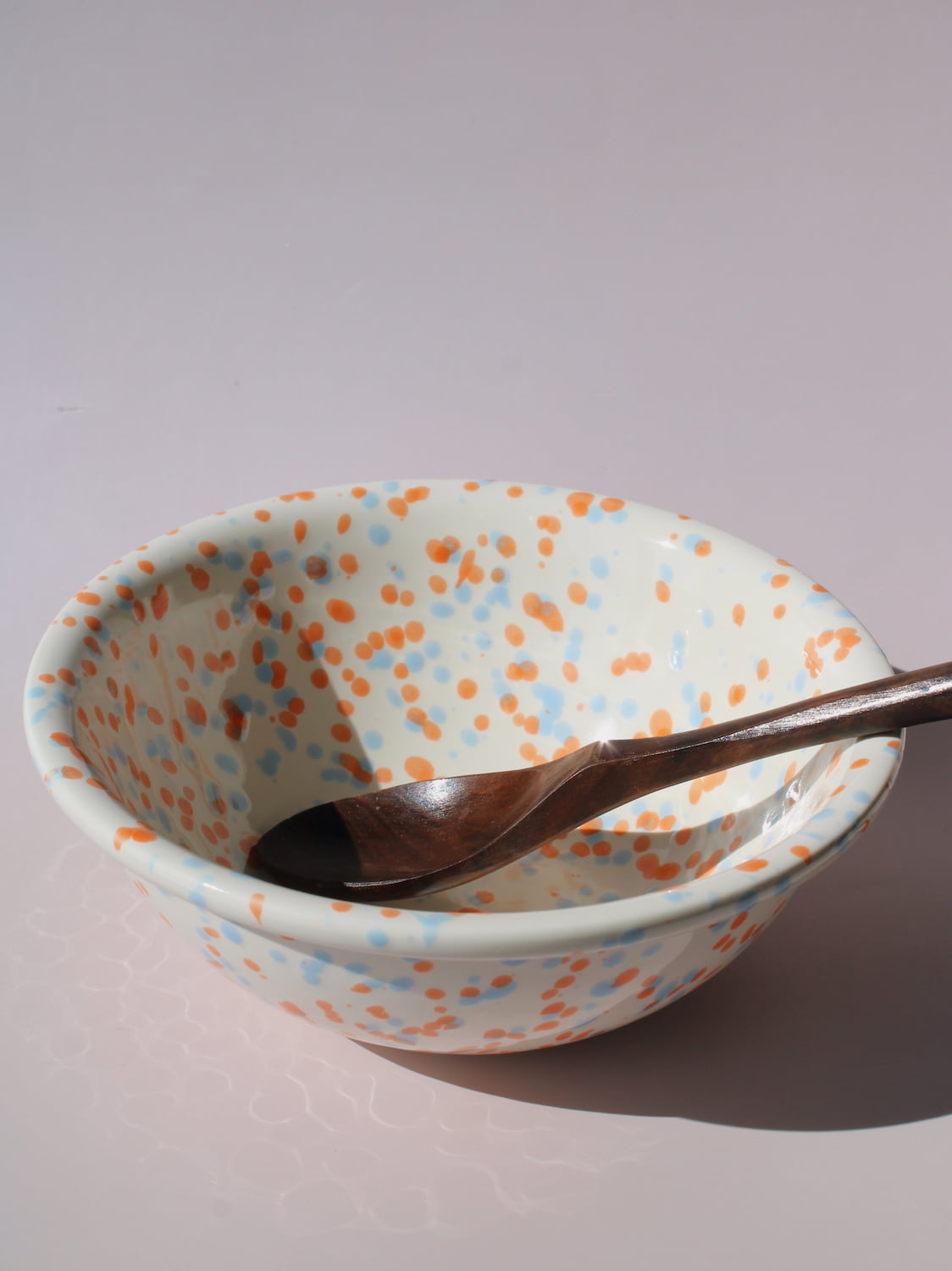 Naro Ceramic Bowl in cream with orange and blue speckled dot pattern, paired with a wooden serving spoon, on a soft pink background.