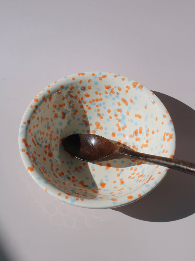 Naro Ceramic Bowl in cream with orange and blue speckled dot pattern, paired with a wooden serving spoon, on a soft pink background.