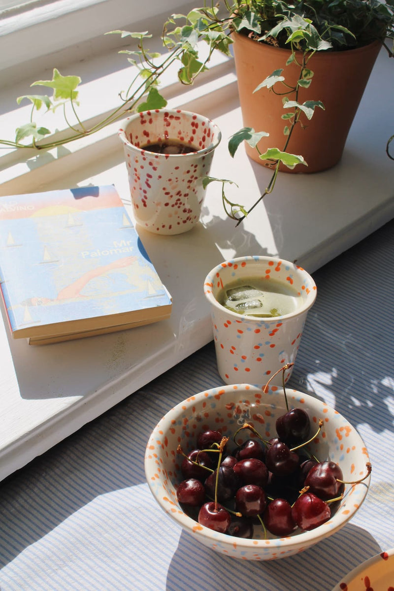 Naro Ceramic Bowl filled with cherries alongside the Naro Ceramic Cup, with the Bera Ceramic Plate with seeded bread and the Bera Ceramic Bowl with raspberries visible on a blue striped surface by a sunny window.
