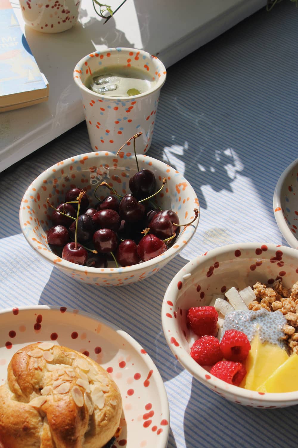 Naro Ceramic Bowl filled with cherries alongside the Naro Ceramic Cup, with the Bera Ceramic Plate with seeded bread and the Bera Ceramic Bowl with raspberries visible on a blue striped surface by a sunny window.