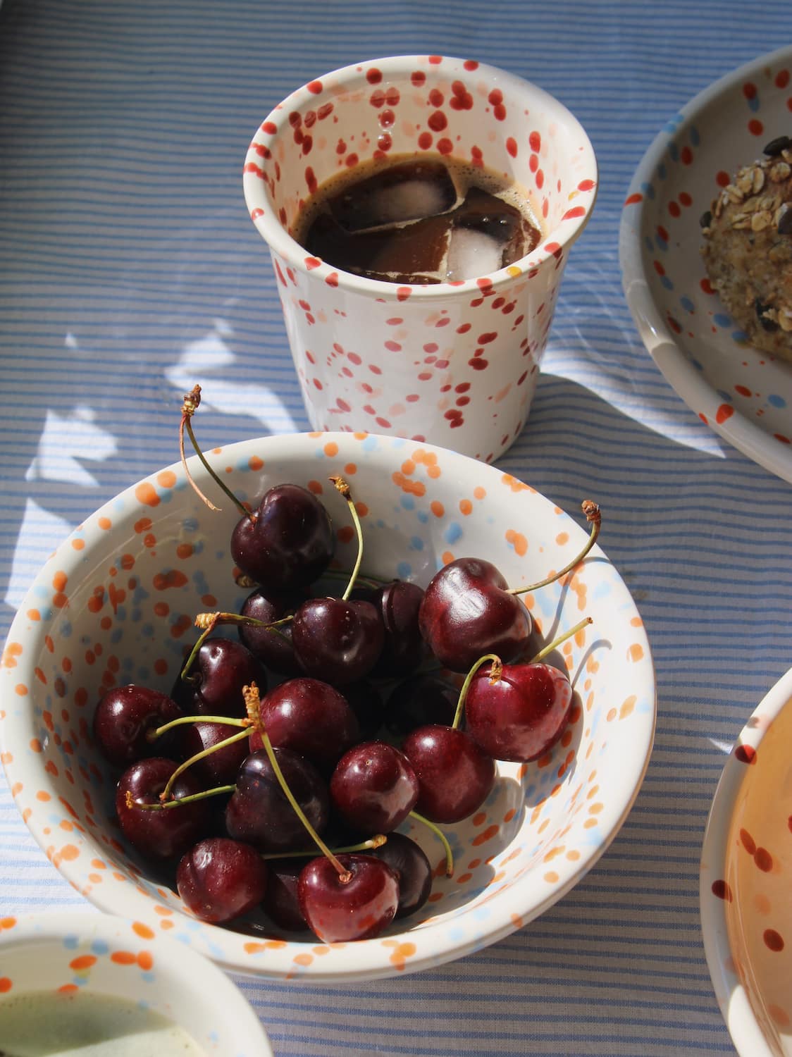 Naro Ceramic Bowl in cream with orange and blue speckled dot pattern, filled with fresh cherries with the Bera Ceramic Cup visible in the background on a blue striped surface in natural sunlight.