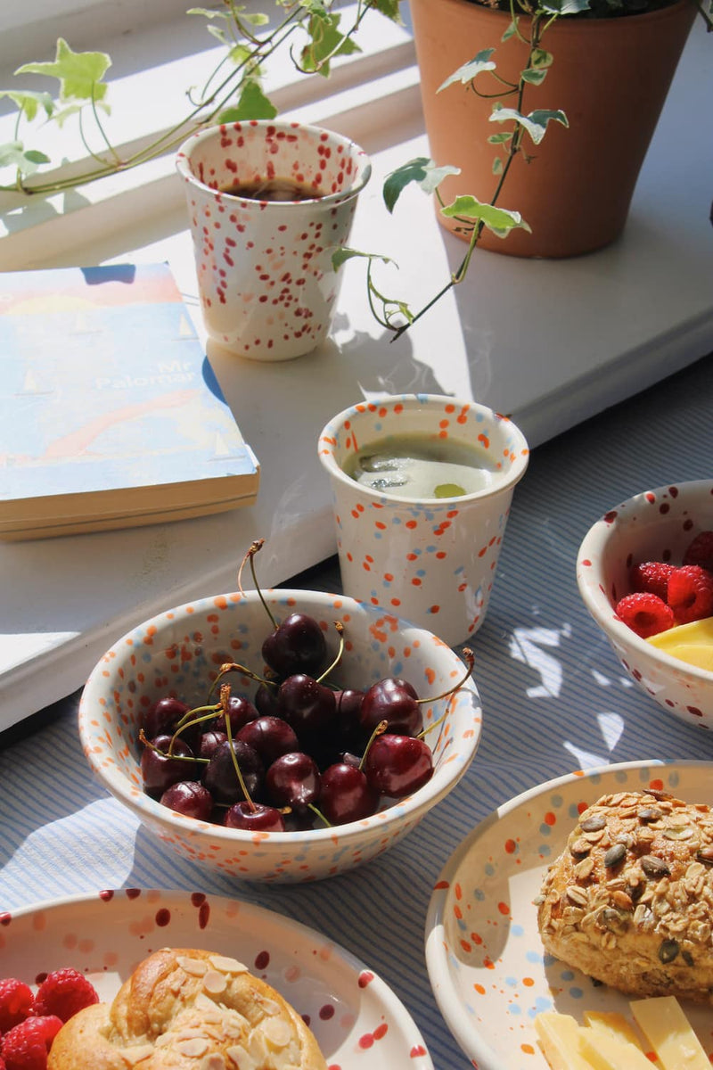 Naro Ceramic Bowl filled with cherries alongside the Naro Ceramic Cup, with the Bera Ceramic Plate with seeded bread and the Bera Ceramic Bowl with raspberries visible on a blue striped surface by a sunny window.