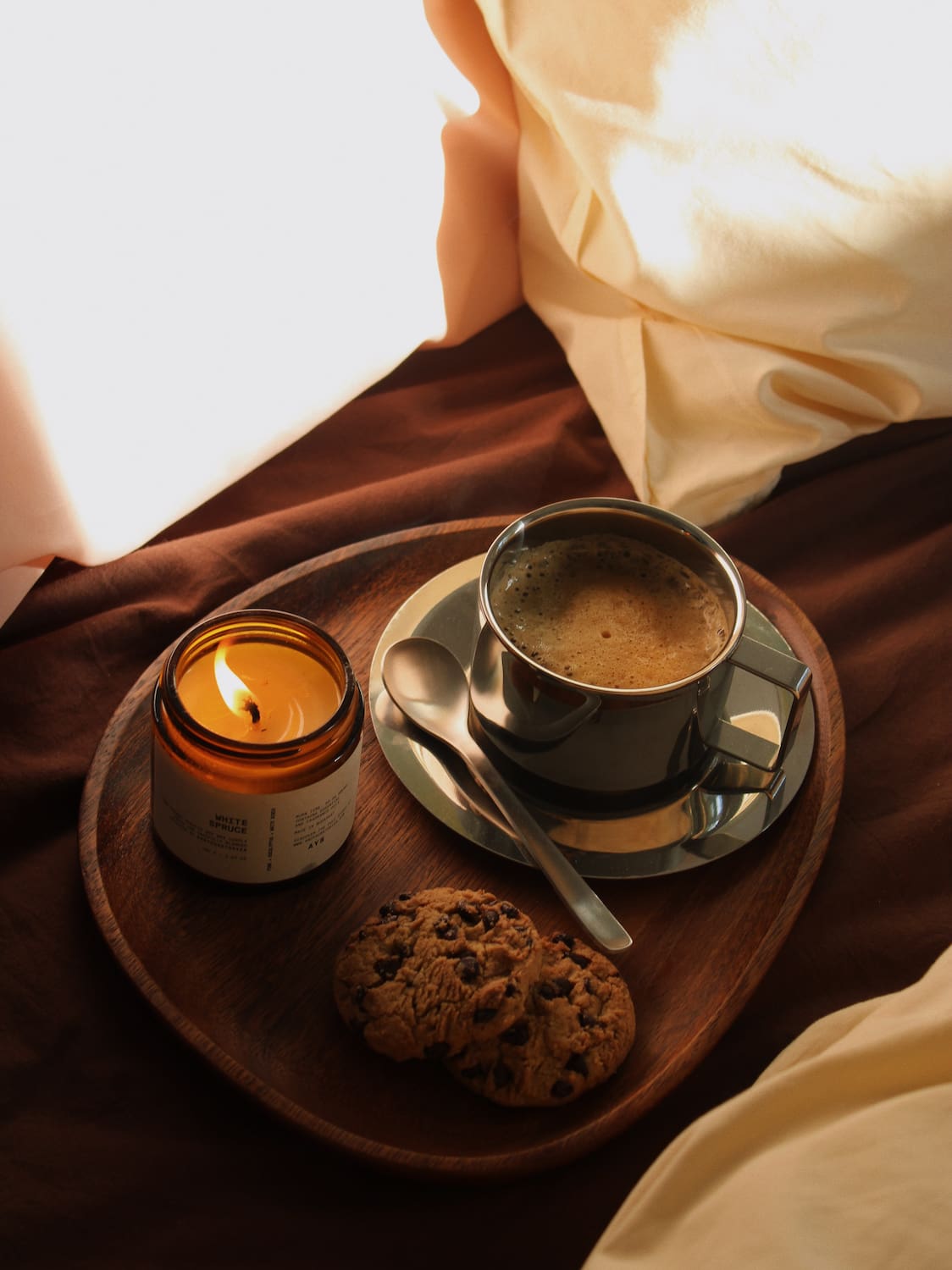 A cozy tabletop scene with a cup of coffee on a saucer, a lit amber candle, a chocolate chip cookie, and a wooden tray resting on dark brown fabric with cream-colored bedding in the background.