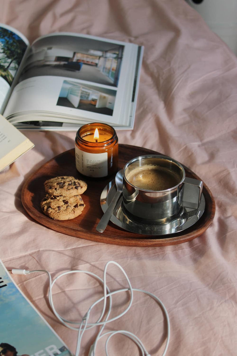 A styled flat-lay scene on pink bedding featuring a wooden tray holding a metal coffee cup with espresso, a lit amber candle, cookies, and a spoon. An open magazine and white earphones are also visible in the scene.