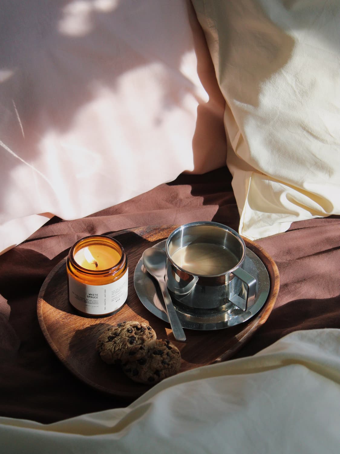 A cozy lifestyle scene featuring a lit amber glass jar candle on a wooden tray next to a stainless steel cup of tea and cookies, set against a background of cream-colored bedding with soft, dappled sunlight.