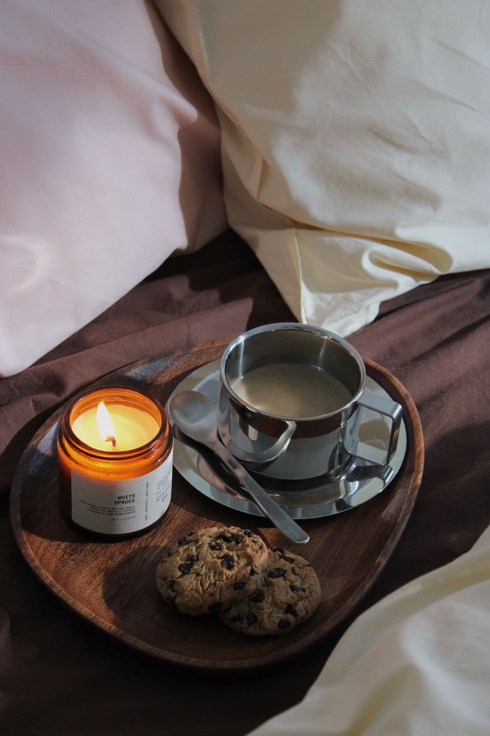 A cozy bedroom scene with a wooden tray holding a lit amber glass candle, a stainless steel mug of coffee on a saucer, a spoon, and two chocolate chip cookies, set against soft pink and cream bedding.