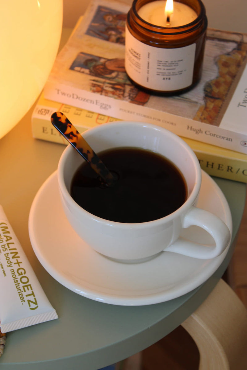A white ceramic coffee cup filled with black coffee sits on a matching saucer on a round table, with a tortoiseshell spoon inside. A lit amber glass candle, books, and a warm glowing lamp are visible in the background.
