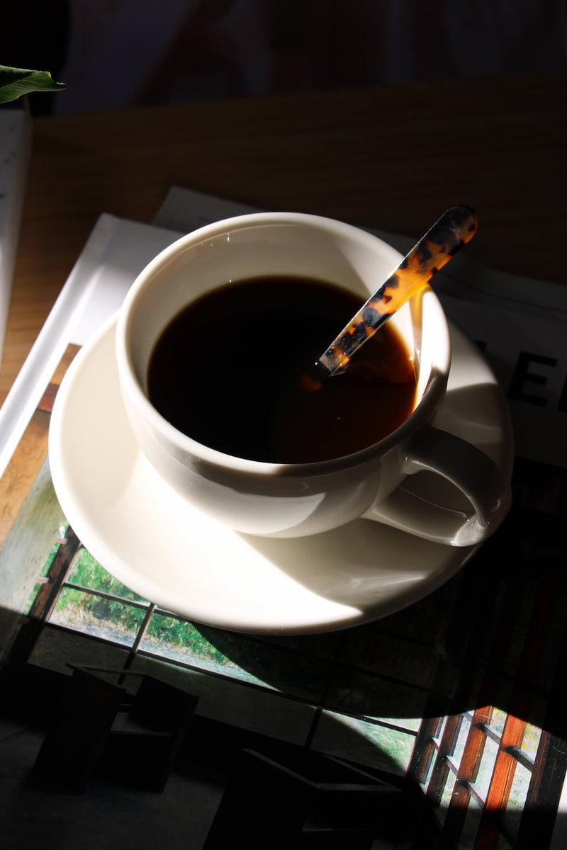 A white ceramic coffee cup filled with black coffee sits on a matching saucer, with a tortoiseshell-patterned spoon resting inside. The cup is placed on top of a book in a setting with strong natural sunlight and deep shadows.