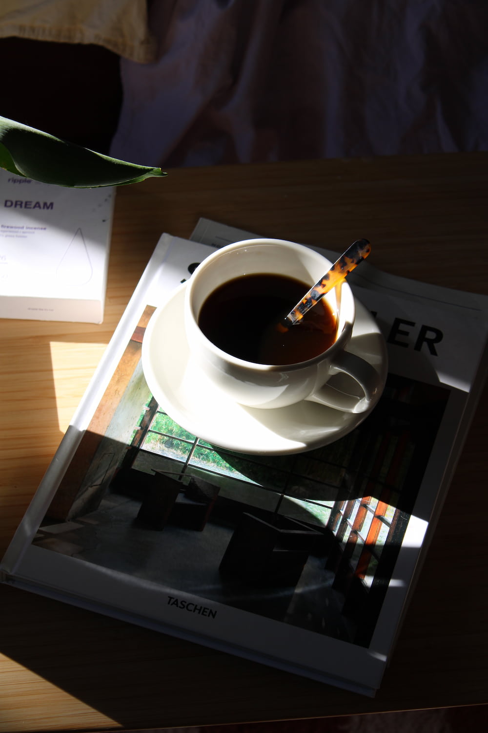 A white ceramic cup of black coffee on a matching saucer, with a patterned spoon resting inside, set on top of a magazine on a wooden table in warm sunlight.