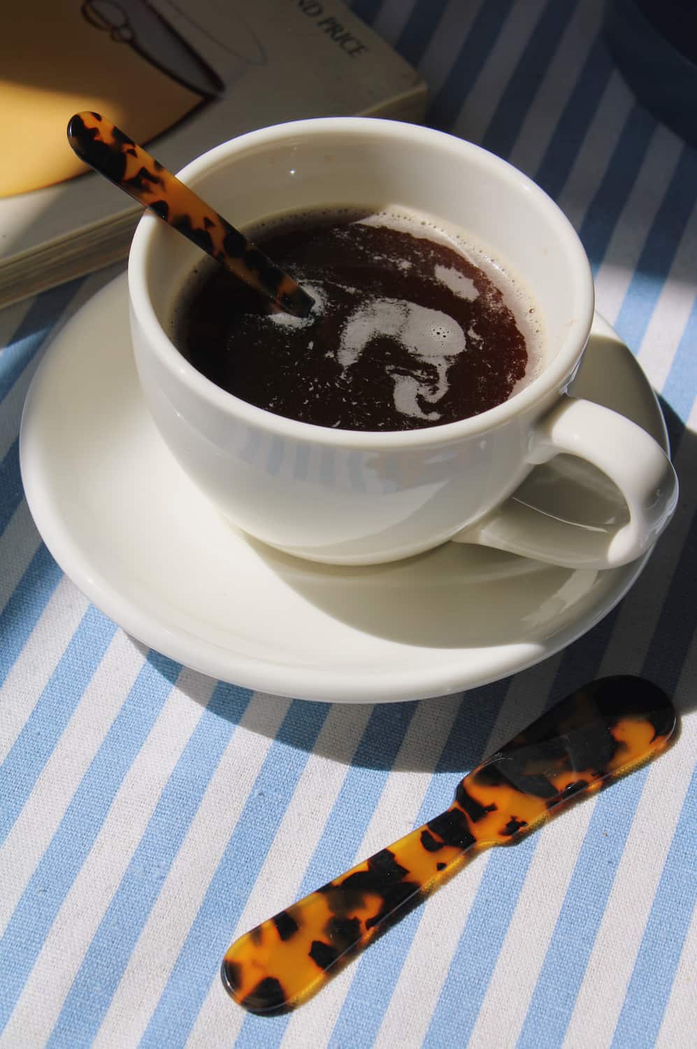 A white ceramic coffee cup filled with dark coffee on a matching saucer, with a tortoiseshell spoon resting beside it on a blue and white striped tablecloth.