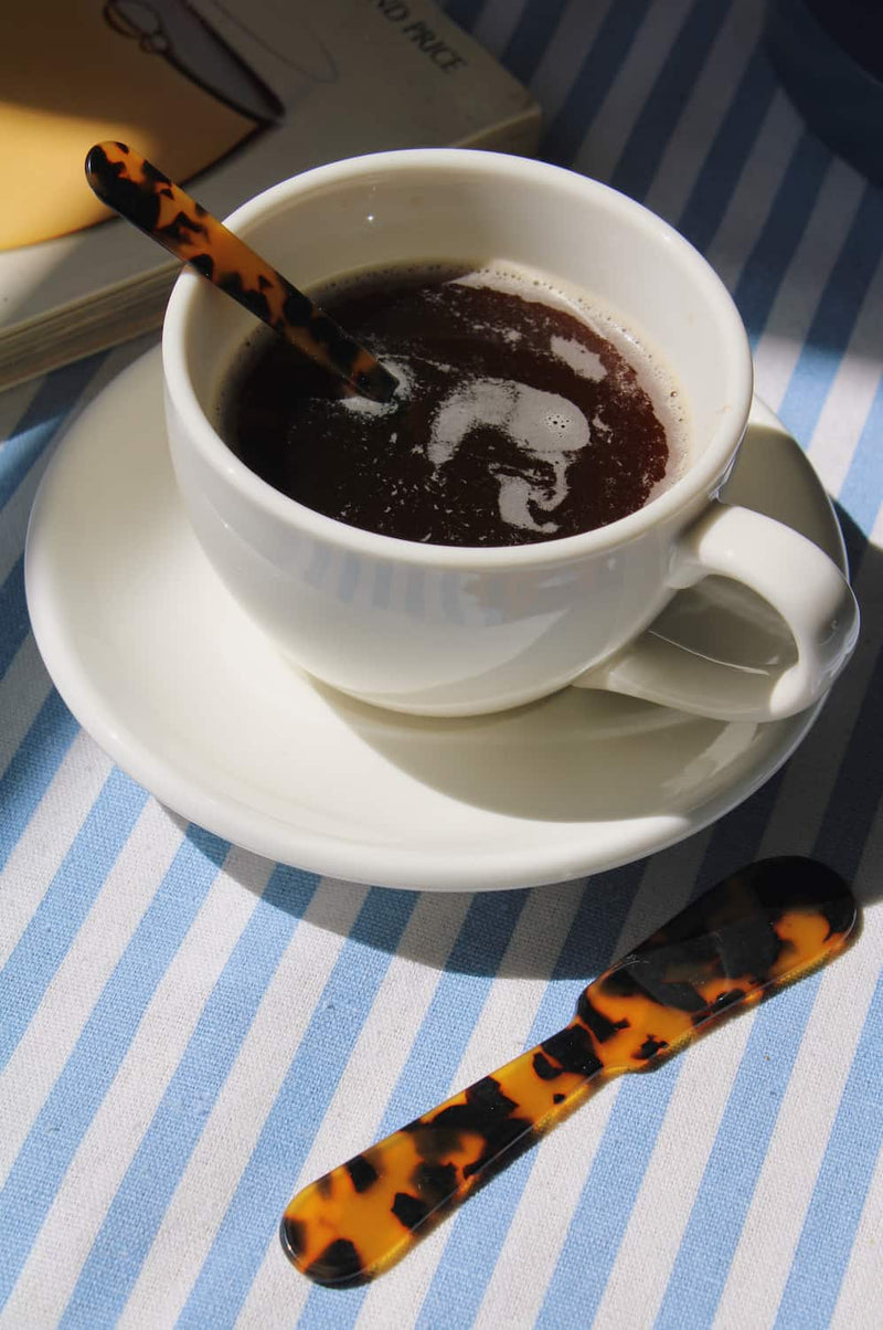 A white ceramic coffee cup filled with dark coffee on a matching saucer, with a tortoiseshell spoon resting beside it on a blue and white striped tablecloth.