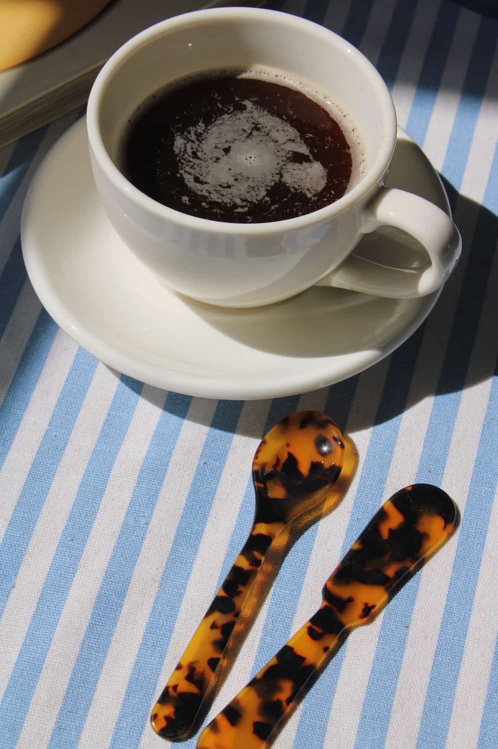 A white ceramic cup of black coffee on a matching saucer, with two tortoiseshell-handled spoons on a blue and white striped tablecloth in bright sunlight.
