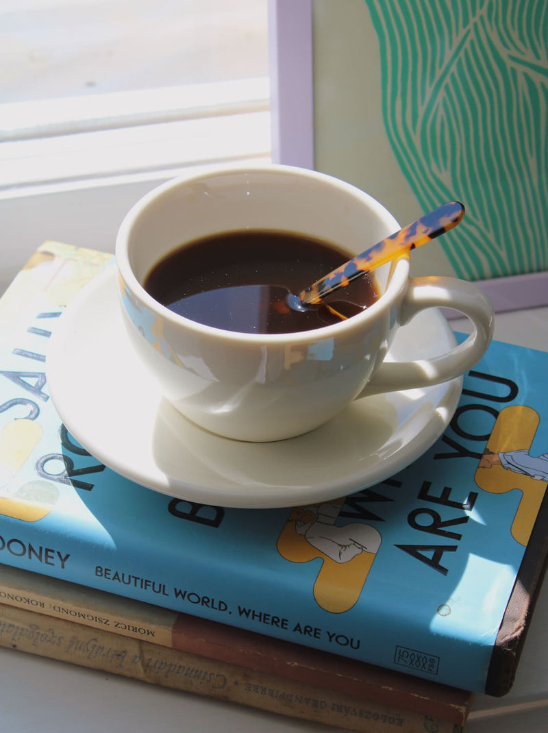 A white ceramic cup of black coffee on a matching saucer, with a patterned spoon resting inside. The cup sits on top of stacked books, including a light blue one with the title 'ARE YOU' visible, near a window with natural light.