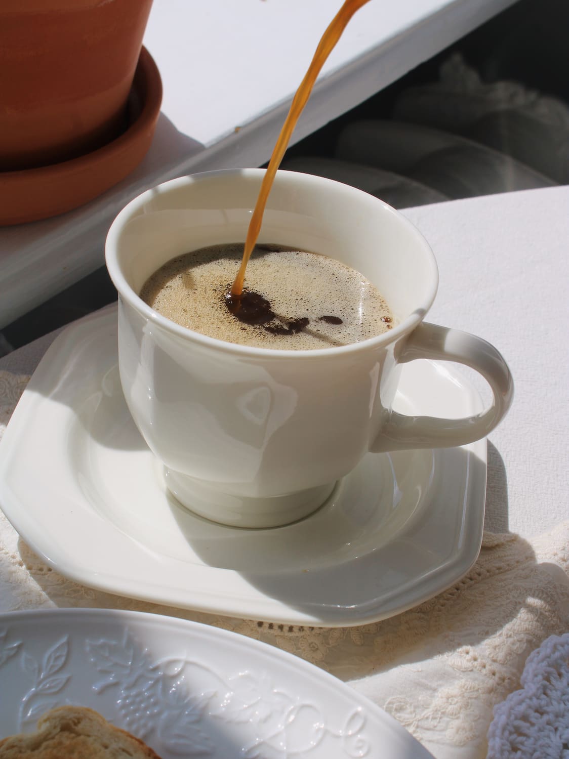 A white ceramic coffee cup on a matching saucer, with coffee being poured into it. The cup and saucer are placed on a white tablecloth, with a terracotta pot in the background and a plate partially visible in the foreground.